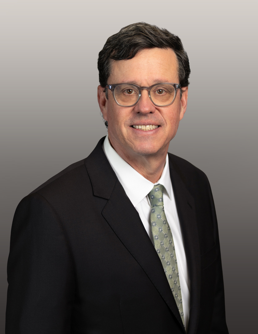 Professional headshot of a man in a dark suit, light shirt, and patterned tie, smiling in front of a neutral background.